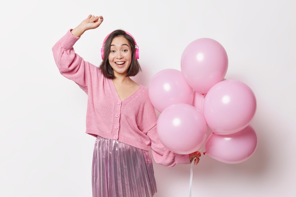 positive-overjoyed-brunette-woman-enjoys-festive-event-holds-bunch-inflated-balloons-anticipates-holiday-dances-carefree-listens-music-via-headphones-wears-jumper-pleated-skirt-poses-indoor